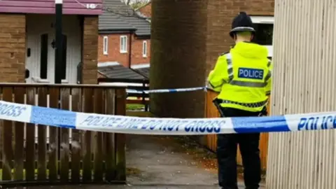 Behind a male police officer outside the houses. A blue and white police cordon is in the foreground.