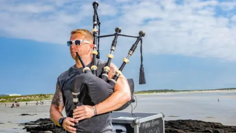 Tiree Music Festival A musician wearing sunglasses and a kilt plays the bagpipes beside the shoreline, with sand, rocks and a cloudy blue sky in the background.