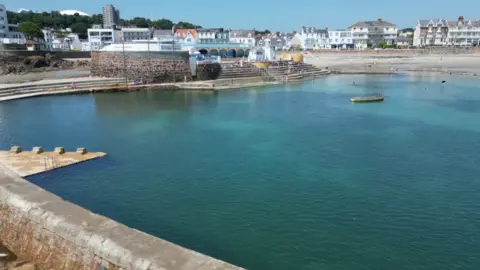 Una gran piscina natural con forma de herradura, llena de agua turquesa, rodeada de muros de adoquines y con casas al fondo.