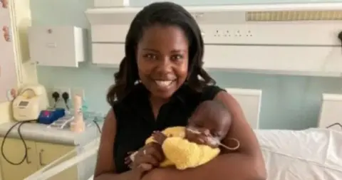 A mother smiles in a hospital ward as she cradles a small baby wearing a yellow knitted cardigan. He has a feeding tube in his nose and his eyes are shut. The mother is wearing a sleeveless black top and has long black hair.