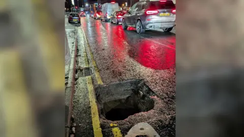 Declan McAteer The sunken manhole on Bridge Street, Newry at night. It has left a gaping hole in the road surface. There are double yellow lines running along the side of the pavement, but part of the lines are missing as the manhole cover has disappeared from view. A line of cars are waiting at traffic lights in the next lane. 
