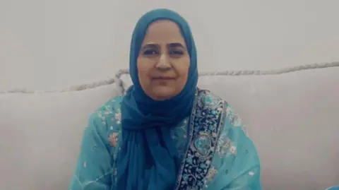 A middle-aged woman wearing a blue dress and hijab sits smiling on a sofa. 