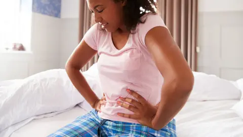 A woman with dark hair sits on a bed and puts her hands on her abdomen. She looks in pain. Shew is wearing blue pyjama trousers and a pink t-shirt