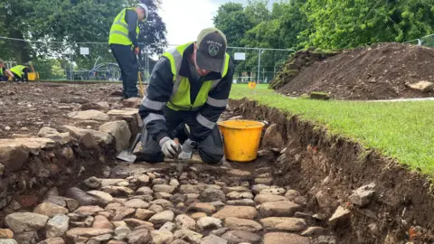 A person wearing high visibility clothing is removing soil from stones in an archaeological site. He is using a small trowel, yellow bucket and black shovel. Behind there is a large pile of soil on green grass and another 3 people also wearing a high visibility vests. There are large trees and a silver fence in the background.