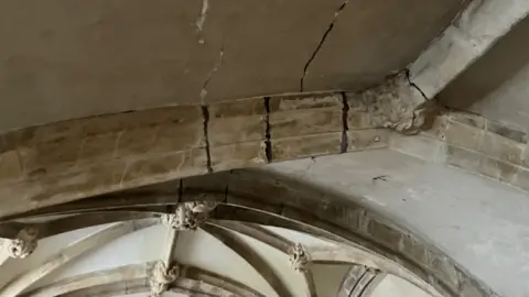 Medieval stone beams show signs of failing historic repair. Several stone ceiling bosses can be seen at the junction of the vaulting. 