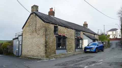 A brown raw brick building with a slate roof and blue car parked outside. 