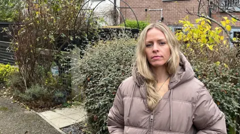 Suzanne Hutton standing in front of a row of houses. She has long blonde hair and is wearing a zipped-up lilac jacket. There are bushes over her shoulder. One of the houses in the background is made from brick, while another has white render.