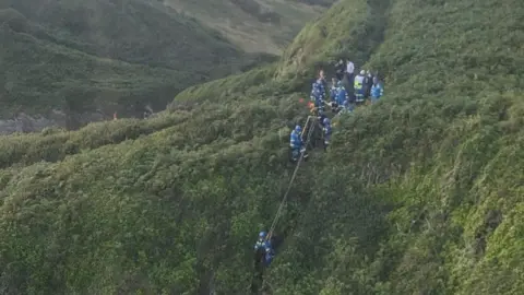 Coastguard teams conduct a rope rescue down the side of a grassy cliff.
