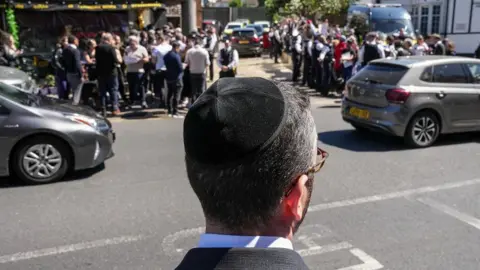 Carlos Jasso/AFP via Getty Images A Jewish man wearing a kippah is seen from behind as he stands looking out at a road as two cars pass in Golders Green. On the opposite pavement crowds are gathered with police officers nearby.