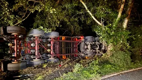 Dorset Roads Policing Underneath and wheels of lorry on its side wedged in undergrowth and trees at the side of a road.