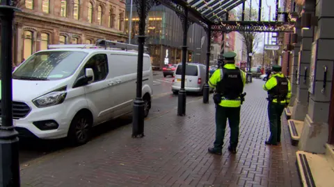 Two police officers on patrol outside Ulster Hall on Belfast's Bedford Street wearing their high-vis, dark green police uniform and black 'police' vest. There are two vans parked on the footpath, one is white and one is grey.