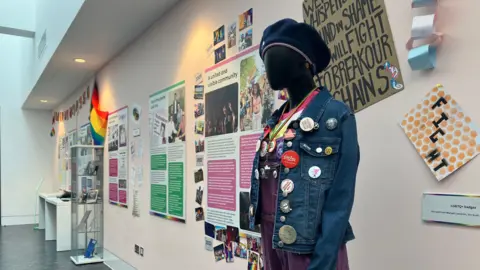 Amy Holmes/BBC A mannequin wearing a badge-covered denim jacket stands beside displays tracing LGBTQ+ history and activism in the “MK Q:munity Tales” exhibition.