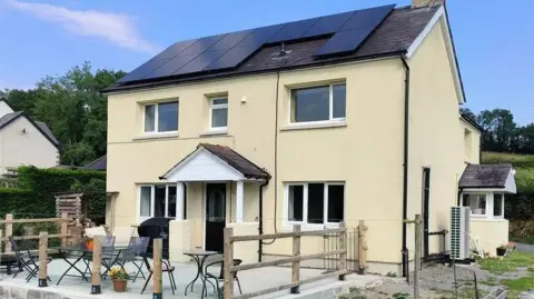 Zoopla/John Francis A two-storey cream-coloured house with a pitched roof fitted with large black solar panels. The house has several white-framed windows, a small covered front porch, and an elevated concrete patio area with outdoor tables, chairs, and wooden railing posts.