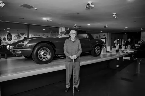 Günter and Christine Starke Ivor Lee stands in front of an E-Type Jaguar. He is wearing a shirt and jumper and is holding a walking stick. 
