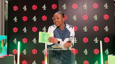 Stacey Olika Olika leaning on a broadcast platform holding Channel 4 que cards in her left hand and a microphone in her right hand, with her forearms arms crossed.
She has her brown hair braided in a parted style and tied back in a bun.
Olika is wearing a blue top with white long sleeve, with blue stars on the shoulders and a gold chain necklace while she lightly smiles at the camera. Behind her is a backscreen covered in the Channel 4 number four logo and 'untold' written in a box style italics.

