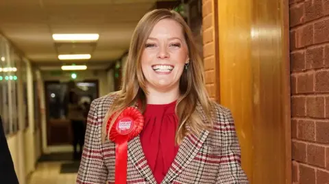 PA Media A woman wearing a red dress and a check blazer walking down a corridor. She is smiling and wearing a red Labour Party rosette on her lapel that reads "Sarah Edwards". She had shoulder length blonde hair.
