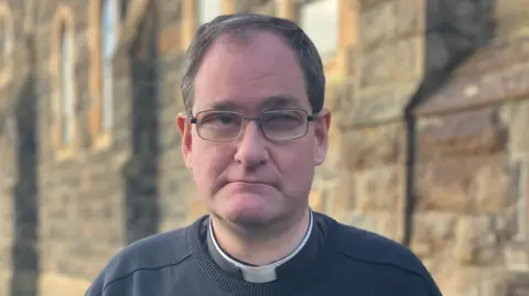 Father Vincent Cushnahan is standing outside a light stone coloured chapel. He is wearing a black jumper with black shirt and white clerical collar. He has dark framed glasses on and short brown hair with some grey hairs. 