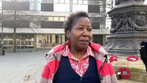 A woman with short black hair is wearing pink earrings, a pink coat, pink checked shirt and navy blue jumper. She is standing by a war memorial with poppy wreaths
