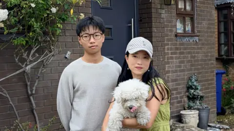 A man in a white jumper, black hair and round rimmed black glasses looks at the camera. Next to him is a woman, smaller than him, wearing a cap and holding a fluffy white dog. They are in front of their home.
