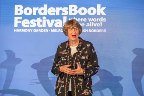 Borders Book Festival Pam Ayres - a woman in a black shirt with pink flowers - stands in front of a blue background