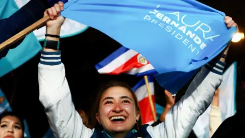 Reuters A supporter of Fernández holds up a flag bearing her name and the word "president" in the light blue colour of her party. The young woman is smiling broadly. Behind her, other supporters can be seen also waving flags. 