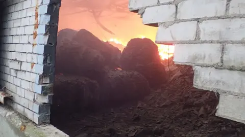 Provided by the Environment Agency Waste on fire seen through a hole in a brick wall on the waste recycling site.