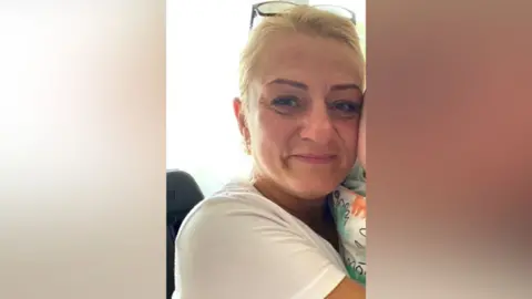 Beata Szauer has blonde hair and is wearing a white shirt while she covers somebody just out of shot.