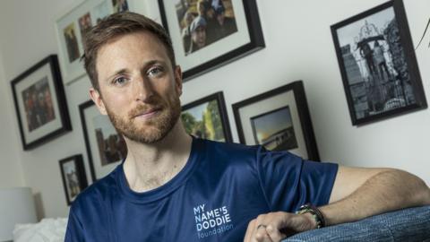 Dr Luke Hames-Brown sitting on a sofa at home - he has short auburn hair and a beard. He is wearing a blue T-shirt and there are family photographs on the wall behind him