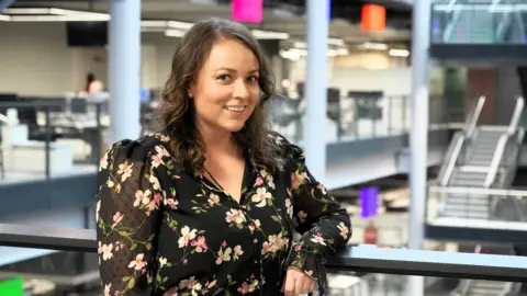 A brunette woman in a dark floral top smiles at the camera as she rests one arm on a bannister in an office 