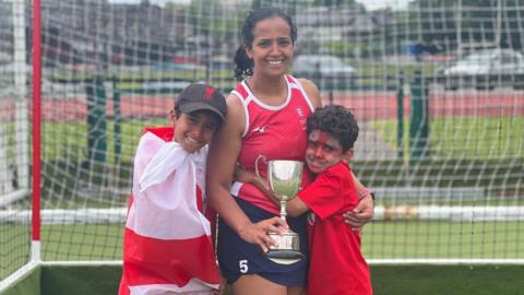 Ms Maan hugging her two sons in front of  hockey goal. she is holding a trophy