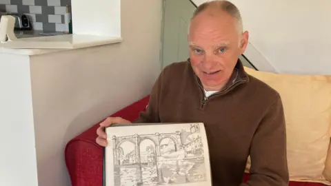 A man is sitting on a red sofa with a yellow cushion behind him holding a hand drawn picture of Knaresborough Viaduct