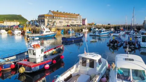 A view across Bridport inner harbour at West Bay on a sunny day. The harbour is full of small colourful boats. An imposing four-storey Victorian terrace sits on the far side of the harbour.