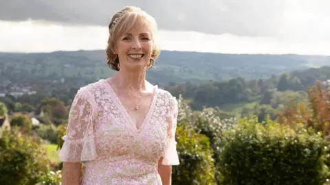 Kate Turner is standing on a hill in a pink dress. She is smiling at the camera as the sun shines on her. She is in a pink lacey and floral dress and has bob-length blonde hair. There is greenery and country hills behind her.
