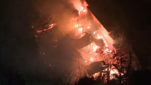 A building covered in fire with its roof collapsed, and otherwise shrouded in darkness, as seen from above. The shadows of some trees can be made out in front of the fire.
