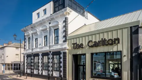 The Carlton cinema seen from the front of the building. The building is made of two main sections. The left side is an 1830s three-storey building painted white with some black detailing, including the sash windows. Around the windows are some decorations such as arches. At the top of the building there is a roof terrace surrounded by a black fence. The section on the right is a metal sage green building on one level, with a tall pitch roof. It has a large window and entrance at the front and the name of the building across the top.