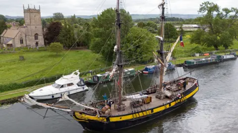 A tall ship is passing through a canal in a countryside area. The ship looks old, is painted black and yellow. It is passing a row of canal boats.