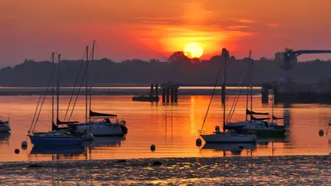Alison Treacher The sun is rising over Gosport harbour. You can see boats silhouetted in the foreground. The sky is glowing orange and reflecting in the water.