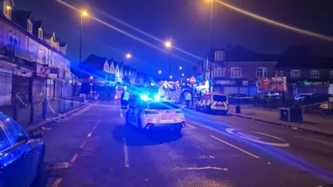 Abdul Khan A police car with blue lights is parked in the middle of the road and there is blue and white police tape visible. It is still dark and the blue lights are reflected across the image.