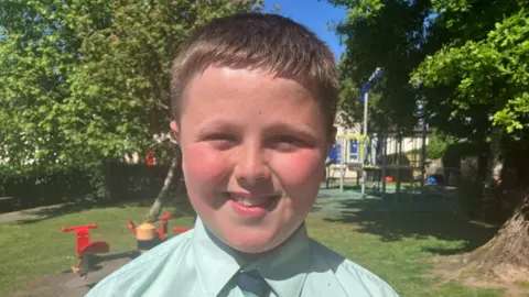 Niall is smiling at the camera. He is wearing a light green shirt and tie. He has short brown hair.