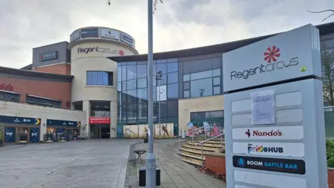 BBC A large shopping complex with a rounded building which says 'Regent Circus' with a glass building to the right and steps.