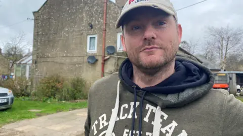 Richard Smith is a man in his 30s wearing a grey top with a cap and some stubble. He is standing in front of a large house with a conservatory just visible to the left