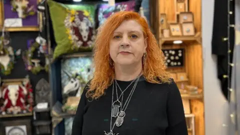 A woman with ginger hair is standing in front of a shop stall and is wearing a black top with metal jewellery.