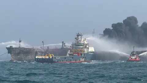 Two ships in the North Sea flanked by a smaller vessel spraying water on them. Thick black smoke can be seen billowing from the vessels in the background 