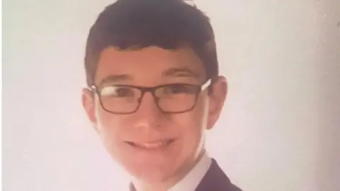 Aaron Webb has short dark hair and black rectangular glasses. He is wearing a white shirt and black blazer.