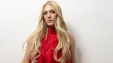 Emily Fern Emily Fern looks directly at the camera while wearing a red dress and posing for a publicity shot against a white background, she has long blonde hair.