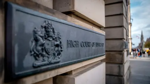 PA Media A general view outside the High Court in Edinburgh. A black sign with dark lettering and a coat of arms is attached to light stone bricks. The lettering reads "High Court of Justiciary". 