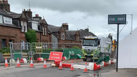 BBC A road closed sign in front of fencing and behind traffic cones next to a sign for a fish and chip shop