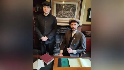 Adam Taylor Alexander Appleton and Danijel Borna Fiket behind a desk at Beamish Museum. Both men are wearing baker boy hats. Danijel is wearing a grey jacket, black waistcoat and a grey sweater. Alexander is dressed in black trousers and a black shirt. There is paper work and a red book on the desk. 