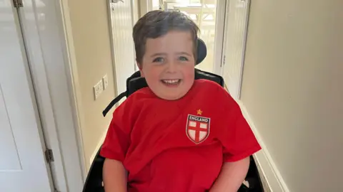 Hannah Gray/BBC A young boy in a red England top sits in a wheelchair in a house.