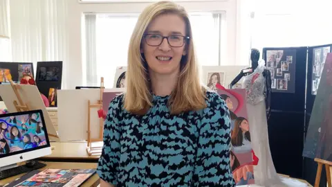 A woman is smiling into the camera. Different art and a mannequin wearing a fancy dress is behind her. She has shoulder-length blond hair, wears glasses and has a blue and black dress on.